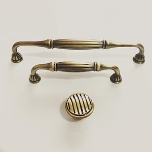 Può includere: Due tiretti per mobili in oro antico con un design scanalato decorativo e un pomo rotondo abbinato con un design scanalato.