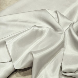 May include: A smooth, white satin fabric with a soft, luxurious drape. The fabric is folded and arranged in a way that shows off its texture and sheen.
