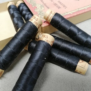 May include: Several black thread spools with paper labels are scattered around a vintage box. The box has German text. The spools are tightly wound, with a glossy finish, indicating a high-quality sewing thread. The labels have text.