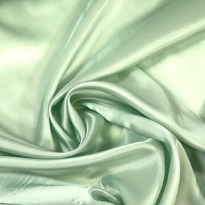 May include: A close-up of a light green satin fabric with a smooth, shiny texture. The fabric is draped and folded, creating soft shadows and highlights. The color is a pale, muted green, reminiscent of seafoam or mint.