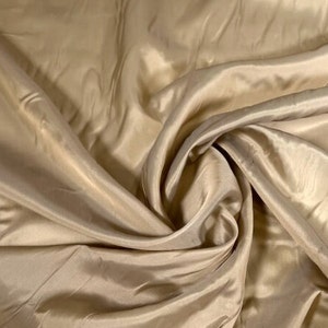 May include: A close-up of a smooth, light beige fabric with a silky texture. The fabric is draped and folded, creating soft curves and folds.