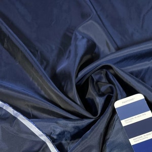 May include: Close-up of a dark blue fabric with a smooth, silky texture. The fabric is draped and folded, showcasing its drape and flow. A color swatch card with the Pantone color names "Ocean" and "Naval Academy" is visible in the bottom right corner.