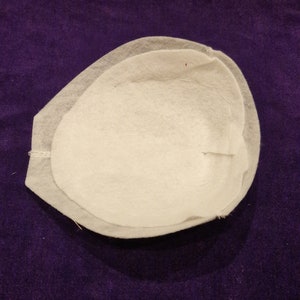 May include: A white fabric cup with a rounded shape and a slightly raised edge. The cup is made of two layers of fabric, with the top layer slightly overlapping the bottom layer.