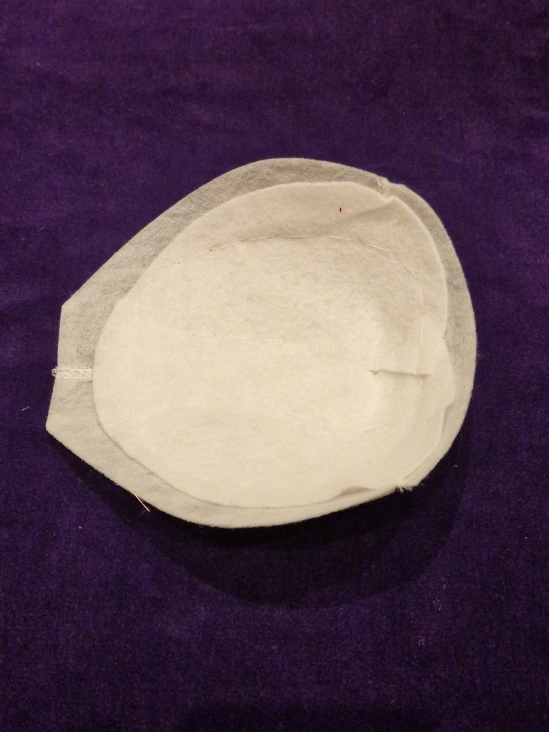 May include: A white fabric cup with a rounded shape and a slightly raised edge. The cup is made of two layers of fabric, with the top layer slightly overlapping the bottom layer.