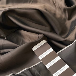 May include: A close-up of a swatch of brown fabric with a silky sheen. A Pantone color card is placed on top, displaying shades of brown, including "Chocolate Brown" and "Delicious". The fabric is draped, showing folds and shadows.