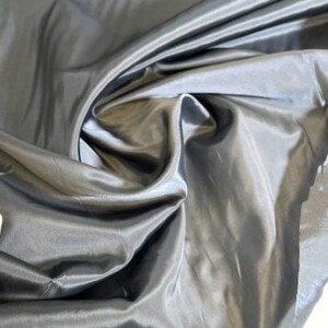 May include: A close-up of a smooth, silver-gray fabric with a silky sheen. The fabric is draped and folded, showcasing its texture and drape.