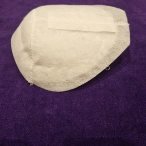 May include: White fabric cup with a stitched seam around the edge. The cup is on a purple surface.