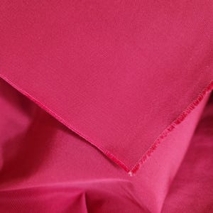 May include: A vibrant magenta fabric featuring a distinct diagonal twill weave pattern. The material is folded to reveal a smooth, matte surface texture with a visible raw edge along the diagonal fold, showcasing the dense and uniform textile construction.