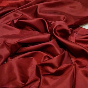 May include: A close-up of a deep red fabric with a smooth, silky texture. The fabric is draped and folded, creating soft, flowing lines.