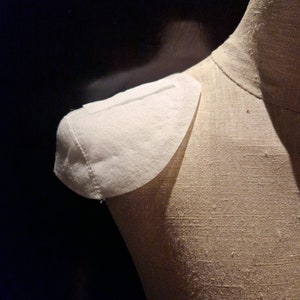 May include: A white fabric collar is pinned to a beige fabric dress form.