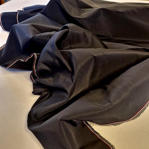 May include: Dark navy fabric with a subtle sheen, draped and folded. The edges are trimmed with a red, white, and blue stripe. The fabric appears to be a medium weight, suitable for apparel or home decor projects.