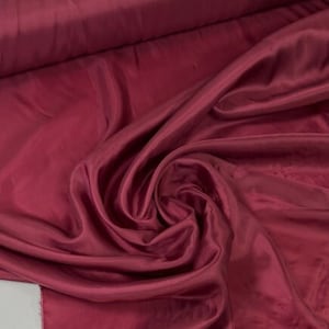 May include: A close-up of a deep red fabric with a smooth, silky texture. The fabric is draped and folded, creating soft curves and folds.