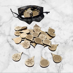 May include: A collection of wooden pendants with engraved geometric designs, including the Flower of Life and Metatron's Cube. The pendants are scattered on a marble surface, with some contained in a black mesh bag.