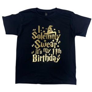 May include: Black t-shirt with gold glitter text that reads "I solemnly swear it's my 11th birthday" with a gold glitter wizard hat graphic.