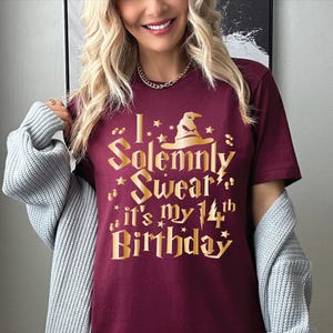 May include: Burgundy t-shirt with gold lettering that reads "I solemnly swear it's my 14th birthday" with a wizard's hat graphic.