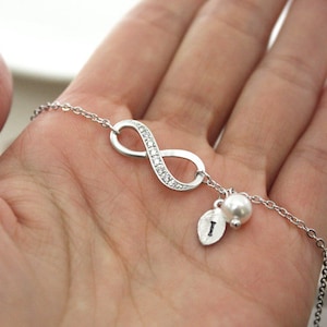 Puede incluir: Un collar de plata con forma de infinito con un pequeño dije de perla y un dije de hoja con la letra 'I'. El símbolo del infinito está incrustado con pequeñas piedras transparentes.