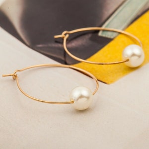 May include: A pair of gold-coloured hoop earrings, each with a large, round, white pearl. The earrings are shown on a cream surface with a yellow and black background. The hoops are thin and delicate.