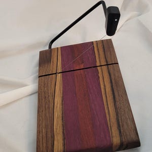 May include: A wooden cheese board with a purple and brown wood grain pattern. A black metal cheese wire cutter is attached to the board.