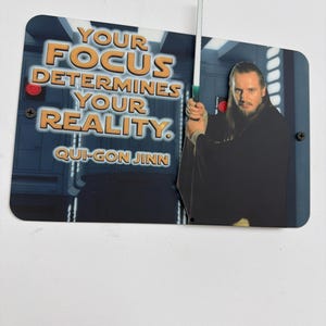 Puede incluir: Un letrero metálico rectangular con Qui-Gon Jinn de Star Wars sosteniendo un sable de luz. El letrero tiene la cita "YOUR FOCUS DETERMINES YOUR REALITY" en texto naranja. El fondo es una escena espacial azul y gris.