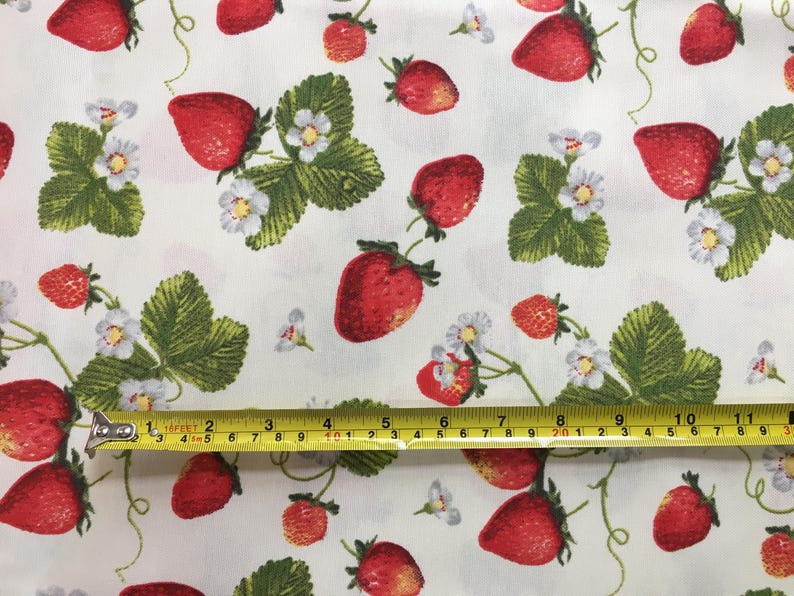 May include: White fabric with a repeating pattern of red strawberries and green leaves. A yellow measuring tape is placed across the fabric, showing measurements in inches.