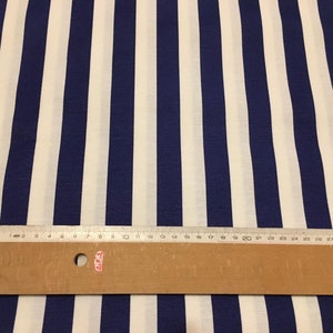 Op de afbeelding: Witte stof met blauwe strepen. Een liniaal ligt op de stof om de breedte van de strepen te laten zien. De liniaal is 76 cm lang.