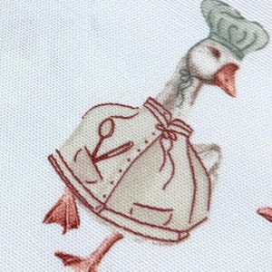 May include: A white goose wearing a chef's hat and a brown apron with a pair of scissors and a spoon embroidered on it. The goose is walking with its legs outstretched.