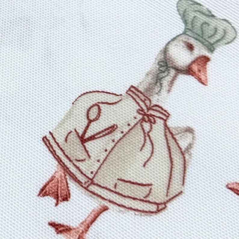 May include: A white goose wearing a chef's hat and a brown apron with a pair of scissors and a spoon embroidered on it. The goose is walking with its legs outstretched.