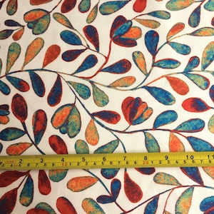 Op de afbeelding: Een witte stof met een herhalend patroon van gestileerde blauwe, oranje en rode bladeren en bloemen. Een gele meetlint ligt over de stof en toont de afmetingen in inches.