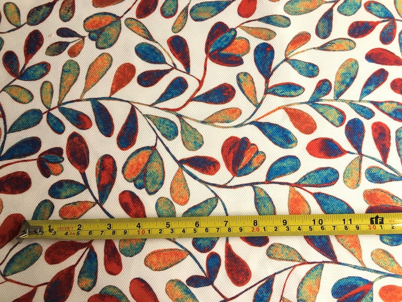 Op de afbeelding: Een witte stof met een herhalend patroon van gestileerde blauwe, oranje en rode bladeren en bloemen. Een gele meetlint ligt over de stof en toont de afmetingen in inches.