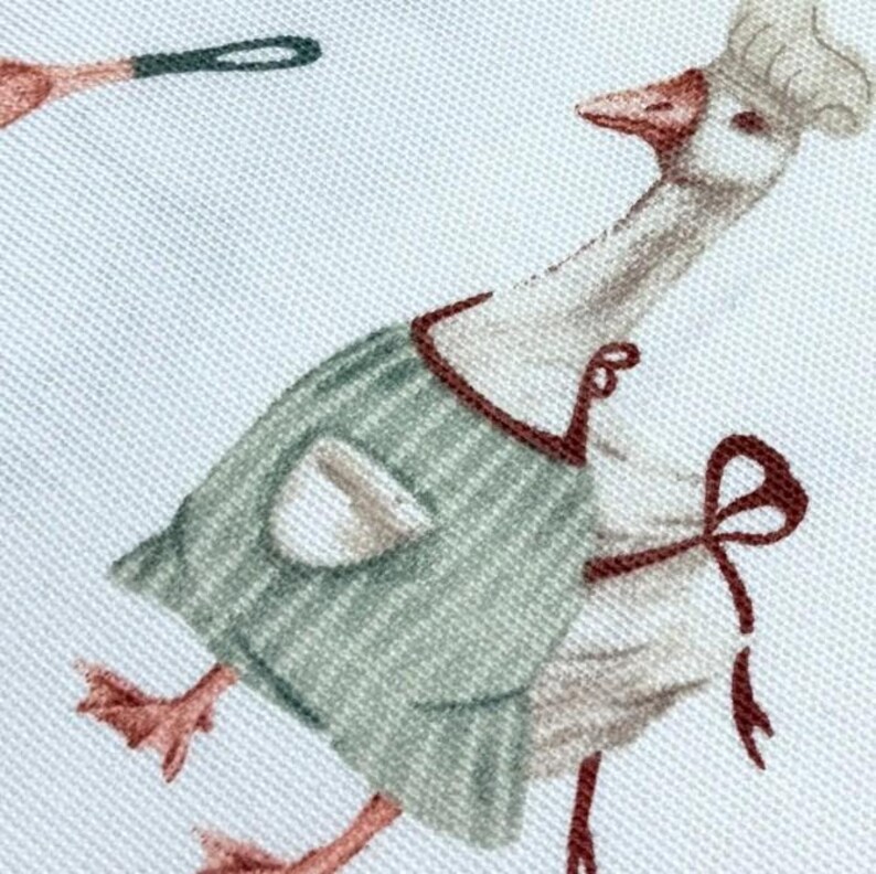 May include: A white goose wearing a green and white striped apron with a brown bow. The goose is wearing a white chef's hat and has a red beak.