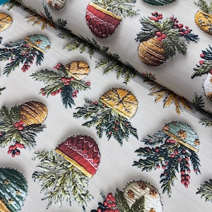 May include: A fabric with a repeating pattern of embroidered Christmas ornaments and evergreen branches. The ornaments are in various colors, including red, gold, green, and blue, with red berries. The background is a light beige.