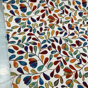 Op de afbeelding: Witte stof met een herhalend patroon van gestileerde blauwe, oranje en rode bladeren. De bladeren zijn donkerblauw omrand en hebben een aquareleffect.