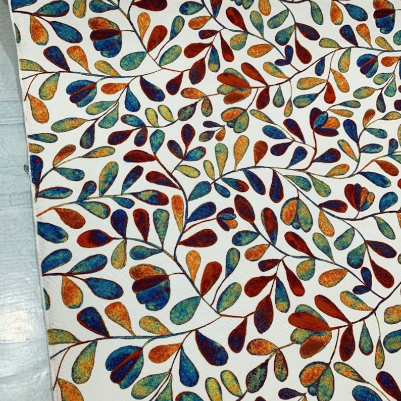 Op de afbeelding: Witte stof met een herhalend patroon van gestileerde blauwe, oranje en rode bladeren. De bladeren zijn donkerblauw omrand en hebben een aquareleffect.