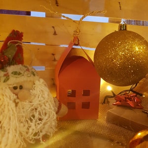 Può includere: Una casetta di carta rossa con finestre ritagliate è appesa a una corda, accanto a una palla di Natale dorata con glitter e una statuetta di Babbo Natale in peluche bianco.