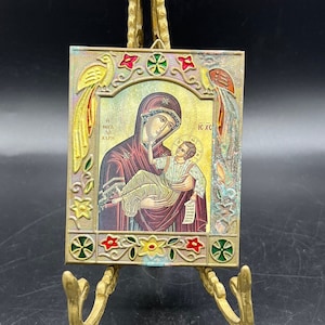 May include: A small, framed religious icon depicting the Virgin Mary holding baby Jesus. The icon is on a gold-colored stand and has a colorful, ornate border with floral designs and birds.