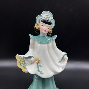 May include: A ceramic figurine of a woman in a white and green dress with a yellow fan. She has black hair and is wearing a white headpiece with green accents.