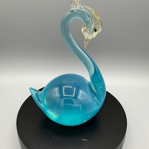May include: A glass swan figurine featuring a vibrant turquoise blue body with a curved neck. The head is crafted from clear glass with subtle gold-toned accents and a small dark eye. The swan is positioned in a graceful, downward-facing pose.