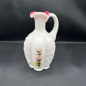 May include: A white milk glass pitcher with a pink rim and floral design. The pitcher has a handle and a bulbous body.