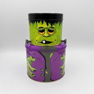 May include: Two Halloween-themed metal tins stacked on top of each other. The top tin is green with a black lid and a Frankenstein monster face. The bottom tin is purple with a green monster hand design.