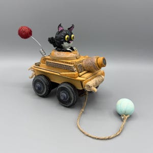 May include: A handcrafted toy tank made of wood and cardboard, featuring a black and white cat figurine on top. The tank has a red yarn ball antenna, a tan barrel, four dark wheels, and a light blue ball attached by a string to the front.