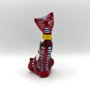 May include: A red ceramic cat figurine with a detailed painted design. The cat has blue and white stripes, a yellow collar, and a detailed face. The figurine is approximately 8 inches tall.