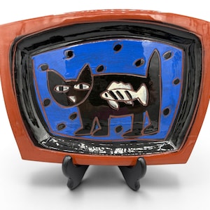 May include: A rectangular ceramic dish with a black cat design holding a fish. The dish has a blue interior with black dots, a black border, and a reddish-brown frame. The dish is displayed on a black stand.