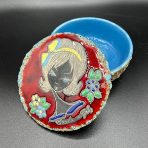 May include: A vintage ceramic trinket box with a lid featuring a stylized portrait of a woman. The lid has a red background with a portrait of a woman with a yellow bow and the text "Carla a Gaby". The box is blue.
