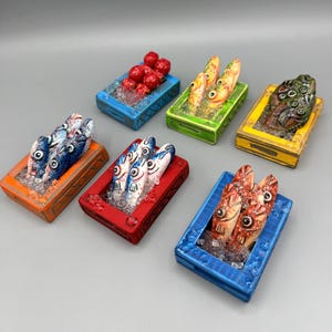 May include: A collection of colorful, miniature fish sculptures displayed in rectangular, open-top containers. Each container is a different color, including blue, red, orange, green, and yellow. The fish have detailed features and are arranged to appear as if they are on ice.