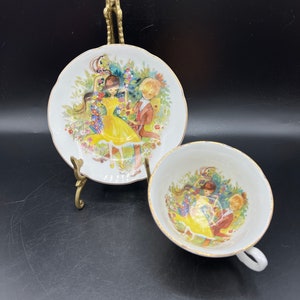 May include: A white teacup and saucer set with a floral design featuring two children dancing in a garden. The saucer has a scalloped edge and the teacup has a gold rim.