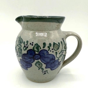 May include: A ceramic pitcher with a gray speckled body, a dark green rim, and a curved handle. The pitcher is decorated with blue floral designs and green leaves.