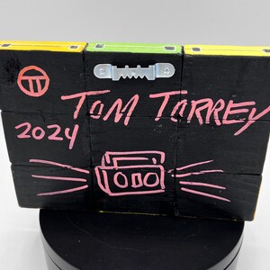 Pu&ograve; includere: Un cartello in legno nero con un cerchio rosa, il testo "TOM TORREY 2024" e un disegno rosa di un boombox con onde sonore che ne escono.
