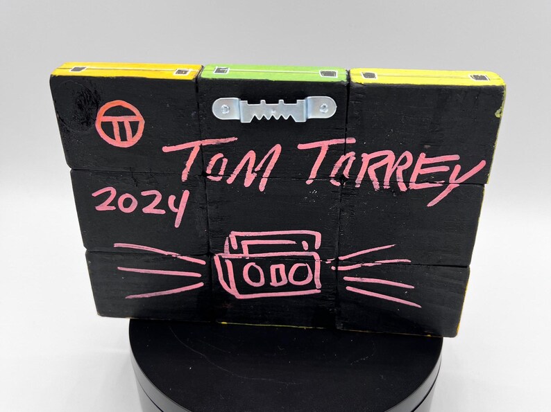 Pu&ograve; includere: Un cartello in legno nero con un cerchio rosa, il testo "TOM TORREY 2024" e un disegno rosa di un boombox con onde sonore che ne escono.