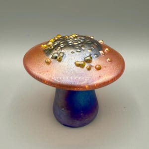 May include: A glass mushroom sculpture featuring a wide, flat cap with a metallic copper and dark gray finish, accented by raised, translucent yellow and amber droplets. The stem is a solid, deep iridescent blue with a smooth, rounded base.