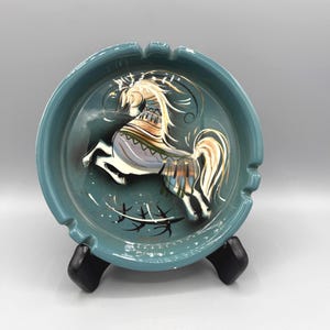 May include: A round teal ceramic ashtray featuring a hand-painted illustration of a rearing horse in shades of white, gold, and black. The horse has a flowing mane and tail with decorative patterns on its body. The ashtray has three notches.